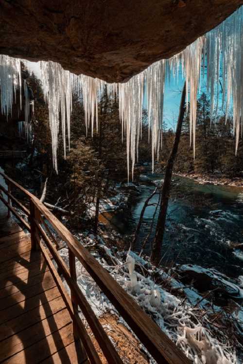 Icicles hang from a rocky overhang above a wooden walkway, with a river and trees visible in the background.