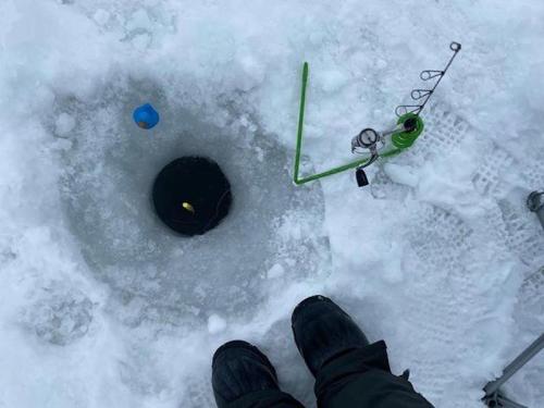A person’s feet in winter boots next to an ice fishing hole, with a fishing rod and bobber visible on the ice.