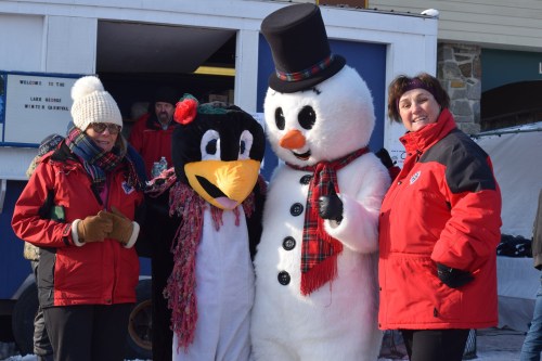 Two women in red jackets pose with a penguin mascot and a snowman mascot at a winter event.