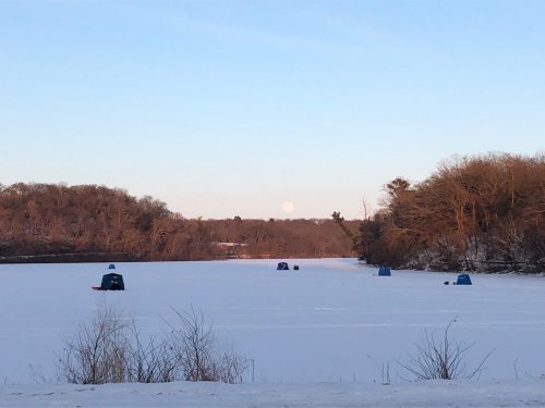 A serene winter scene with ice fishing huts on a frozen lake, under a clear sky with a rising moon in the background.
