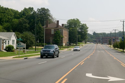 A quiet street scene with a few cars, trees, and buildings lining the road on a sunny day.