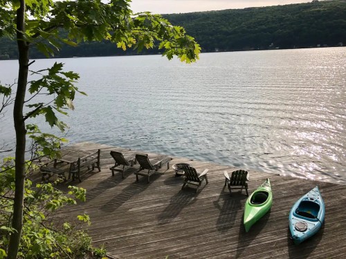 A serene lakeside scene with two kayaks and wooden chairs on a dock, surrounded by trees and calm water.