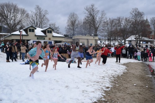 A group of people in swimwear runs into a snowy beach while a crowd watches from the shore.