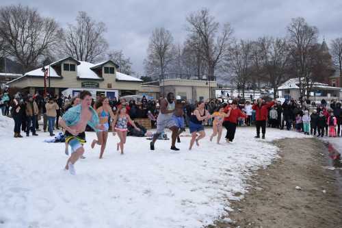 A group of people in swimwear runs into a snowy beach while a crowd watches from the shore.