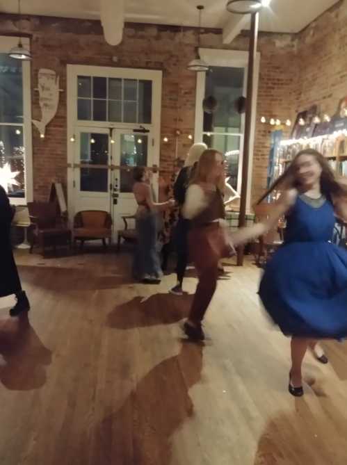 A lively indoor scene with people dancing in a warmly lit space, featuring a mix of dresses and a rustic atmosphere.