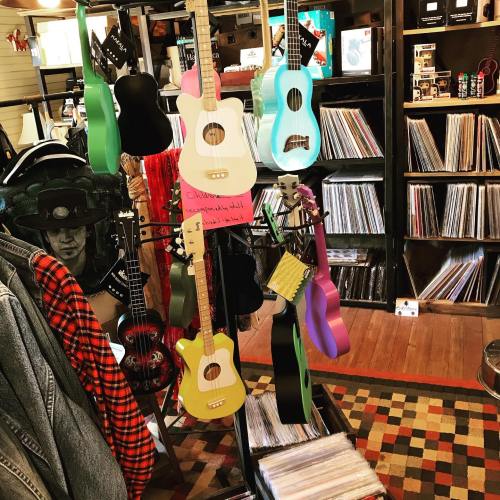 A colorful display of guitars and ukuleles in a music store, with vinyl records visible in the background.