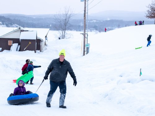 A man pulls a child on a sled through snowy terrain, while others enjoy sledding in the background.
