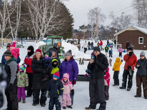 A crowd of people, including children in winter clothing, gathers outdoors in a snowy area with a tractor and buildings in the background.