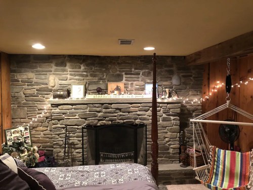 Cozy basement with a stone wall, fireplace, decorative items, and a hammock chair, illuminated by string lights.