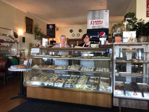 Two bakers stand behind a display case filled with various baked goods in a cozy bakery setting.