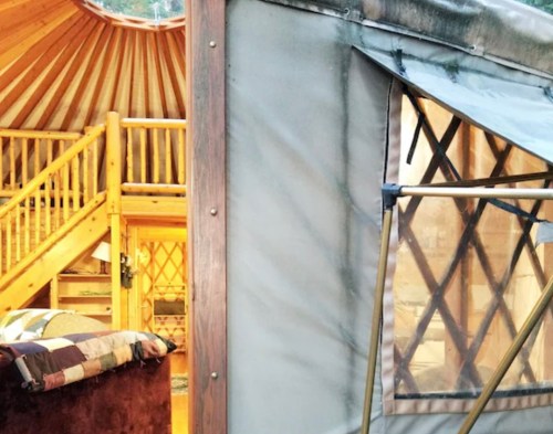 Interior of a cozy yurt with wooden beams, a staircase, and a view of a quilted bed through a window.