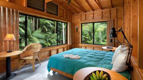 Cozy wooden bedroom with a turquoise bedspread, large windows overlooking lush greenery, and a small table with a lamp.