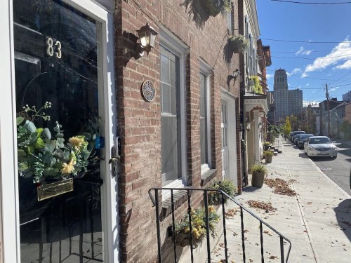 A brick building with a decorative door, plants, and a sidewalk lined with autumn leaves and parked cars.
