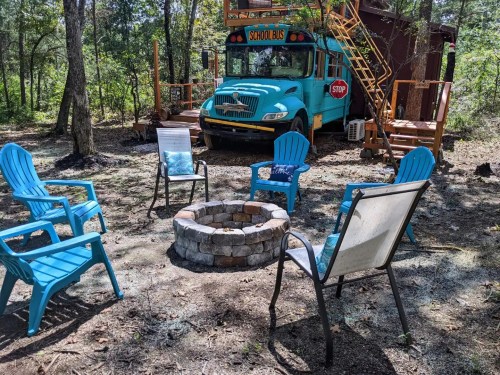 A colorful school bus converted into a living space, surrounded by chairs and a stone fire pit in a wooded area.
