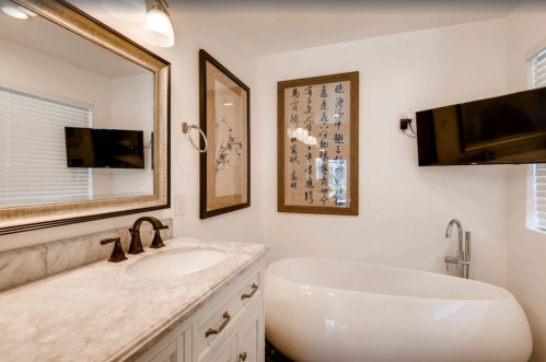 Modern bathroom featuring a freestanding tub, marble countertop, large mirror, and wall art. Two TVs mounted on the walls.
