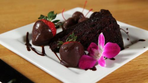 A slice of chocolate cake garnished with chocolate-covered strawberries and a pink orchid on a white plate.