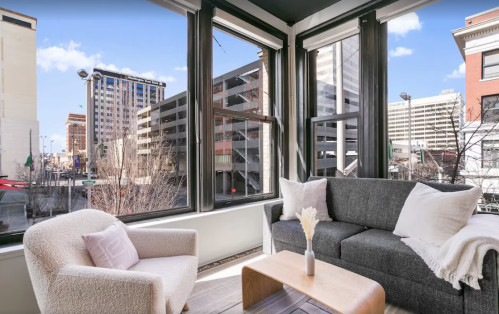 Bright, modern living room with large windows showcasing a city view, featuring a gray sofa and a cozy armchair.