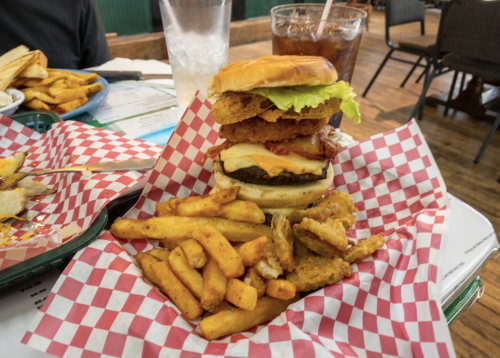 A stacked burger with fried chicken, lettuce, and cheese, served with fries in a red-checkered basket.