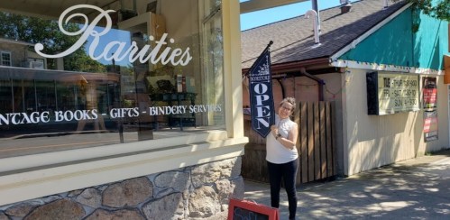 A woman stands outside a shop called "Rarities," holding an "Open" flag in front of a vintage bookstore.