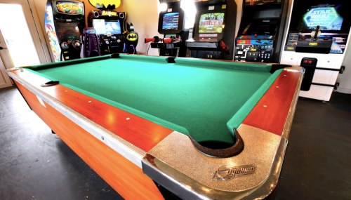A green pool table in a game room with arcade machines in the background. Brightly lit and inviting atmosphere.