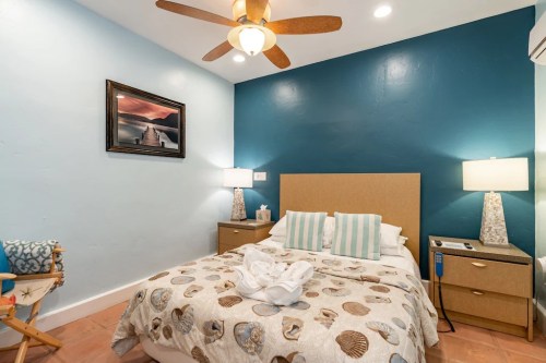 Cozy bedroom with a blue accent wall, a queen bed, nightstands, lamps, and a ceiling fan. Decorated with seashells.