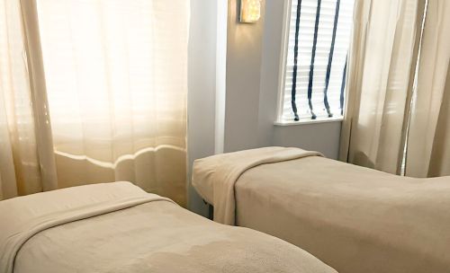 A serene spa room with two massage tables, soft lighting, and sheer curtains filtering natural light.
