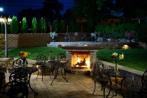 A cozy outdoor patio at night, featuring a stone fireplace, tables with flowers, and lush greenery in the background.
