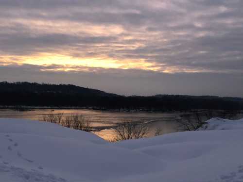 A serene winter landscape with snow-covered hills and a river reflecting a cloudy sunset.