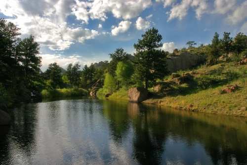 A serene landscape featuring a calm pond surrounded by trees and rocky hills under a partly cloudy sky.