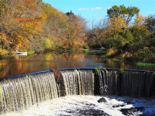 A serene waterfall cascades into a calm river, surrounded by vibrant autumn foliage and clear blue skies.
