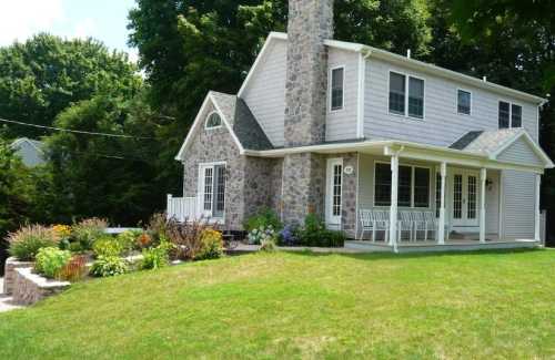 A two-story house with a stone chimney, surrounded by a lush garden and green lawn on a sunny day.