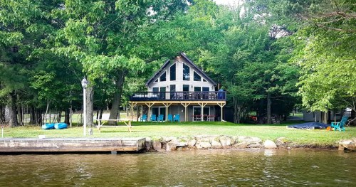 A cozy cabin by the lake, surrounded by trees, with a spacious deck and colorful chairs on the lawn.