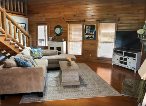 Cozy living room with wooden walls, a sectional sofa, coffee table, and a TV, featuring large windows with blinds.