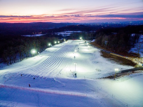 A snowy landscape at dusk, featuring illuminated ski slopes and a serene sunset in the background.