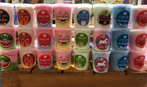 Colorful tubs of cotton candy in various flavors stacked on a wooden display, each labeled with vibrant designs.