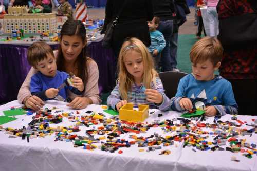 A woman and three children play with colorful LEGO bricks on a table at a busy event.