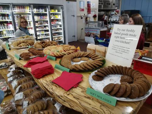 A display of various freshly baked cookies on a wooden table, with shoppers in the background and a sign promoting the goods.