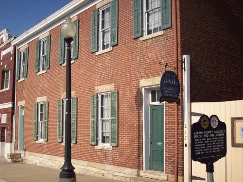 Historic brick building with green shutters, a sign reading "Jail," and a nearby informational marker.