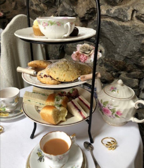 A tiered stand with assorted pastries, sandwiches, and a teacup, set against a stone wall backdrop.