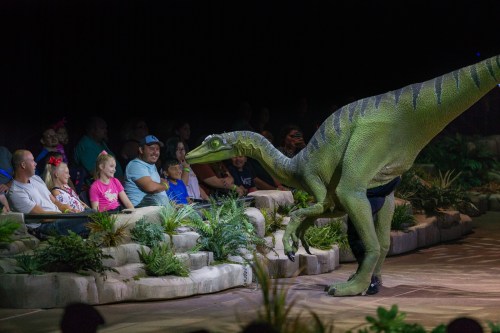 A lifelike dinosaur puppet interacts with an audience of families in a lively performance setting.
