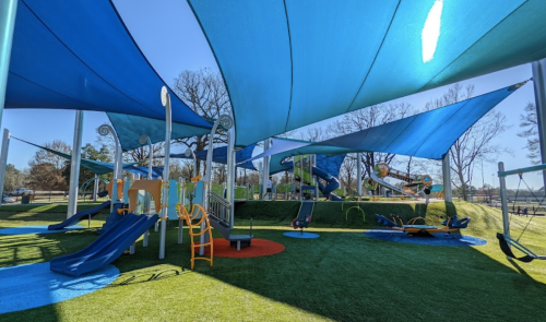 A colorful playground with blue shade sails, slides, swings, and climbing structures on green turf.