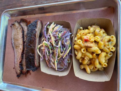 A tray with smoked brisket, coleslaw, and macaroni and cheese served in paper containers.