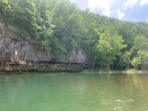 A serene river scene with lush green trees and rocky cliffs under a partly cloudy sky.