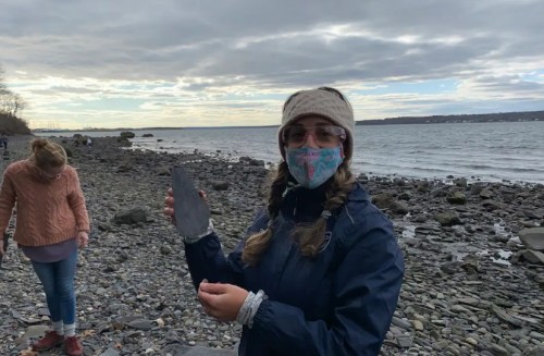 A person wearing a mask and winter clothing holds a slate rock by the shore, with another person collecting stones nearby.