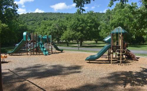 A playground with slides and climbing structures, surrounded by trees and a grassy area under a blue sky.