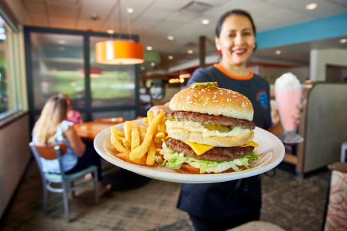 A server holds a plate with a large burger and fries, with a milkshake in the background and diners at a table.