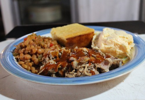 A plate of pulled pork with barbecue sauce, baked beans, cornbread, and creamy mashed potatoes.