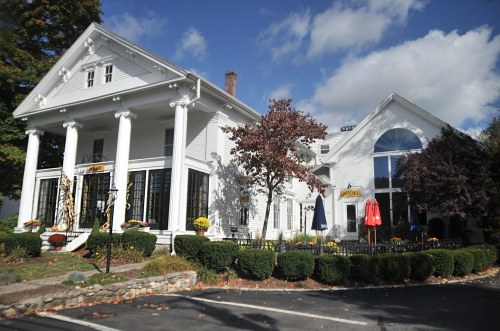 A large white house with columns, surrounded by greenery and autumn decorations, under a blue sky with clouds.