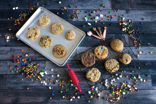A variety of cookies on a baking tray and cooling rack, surrounded by colorful sprinkles and baking tools.
