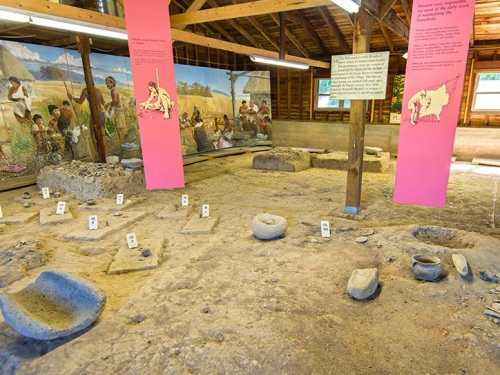 Archaeological site with stone tools and artifacts, surrounded by informative panels and murals depicting historical scenes.